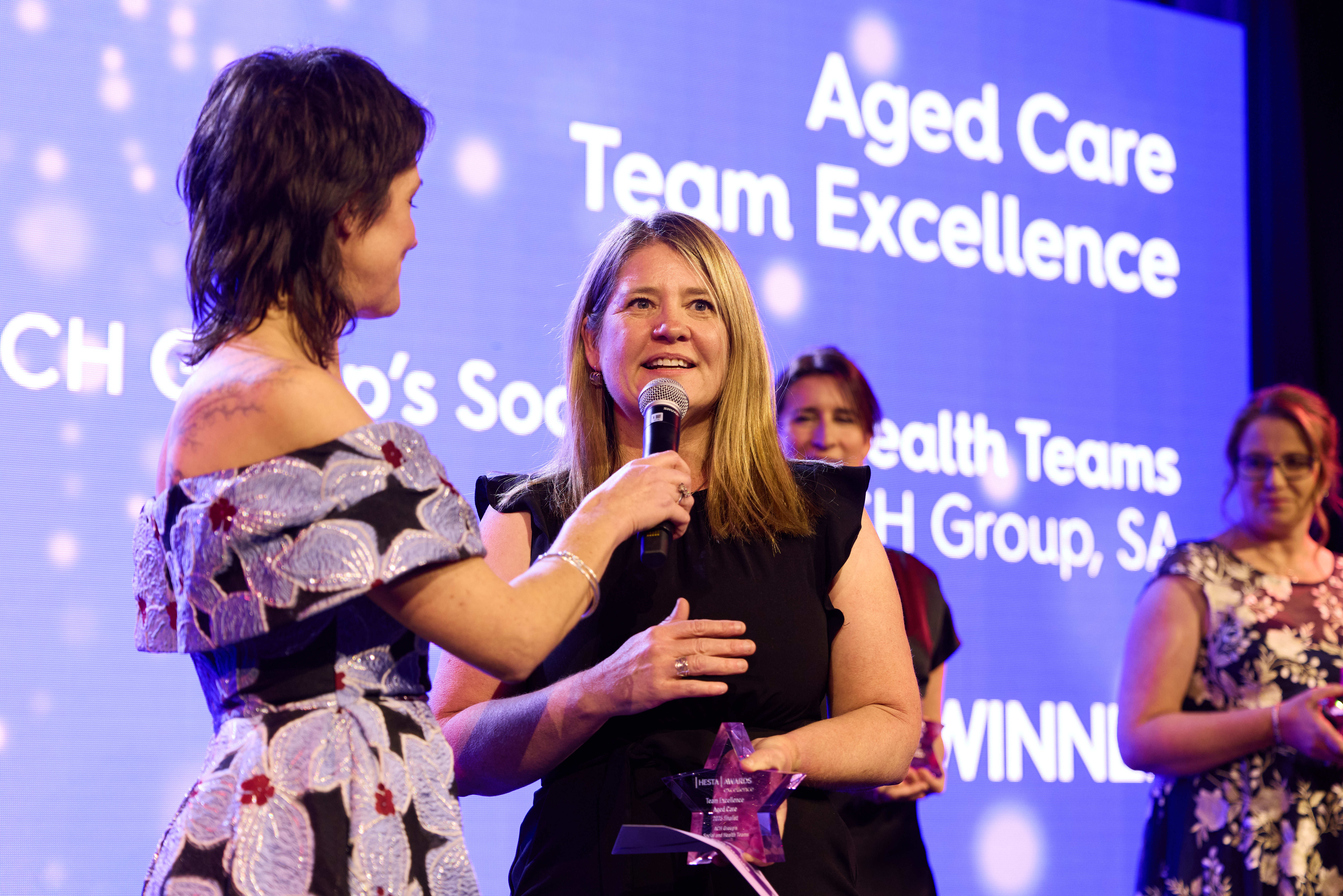 A female HESTA Aged Care Team Excellence Awards winner is being interviewed on the stage.