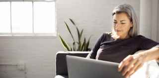 Image of a woman with grey hair sitting on a couch smiling and using a laptop.